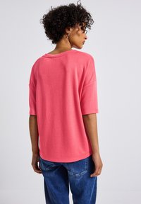 Korálově růžové oversized tričko s širokým výstřihem a krátkými rukávy, vyrobené z měkké tkaniny. Kombinováno s modrými volnými džíny.