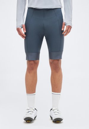 Άνδρας που φοράει σκούρα γκρι ποδηλατικά σορτς, λευκό μακρυμάνικο μπλουζάκι, λευκές κάλτσες και λευκά ποδηλατικά παπούτσια σε απλό φόντο.