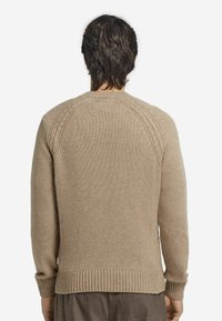 Beige stickad tröja med ribbad fåll och manschetter, med texturerat axeldetalj, visad bakifrån.