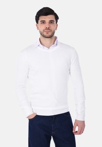 Maglione bianco a V in morbido tessuto con maniche lunghe, abbinato a una camicia colletto blu chiaro. Jeans con una texture di denim scuro completano l'outfit.