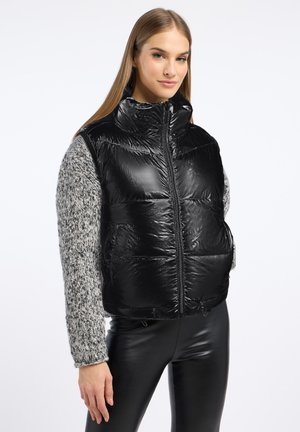 Zwarte puffer vest met hoge kraag, glanzende afwerking, voorrits en elastische trekkoordzoom, gecombineerd met een grijze gebreide trui met lange mouwen.