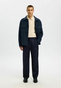 Plava jakna od vune s dva prednja džepa, bež polo majica, mornarsko plave hlače i crne cipele. Model stoji nasuprot jednobojnoj pozadini.