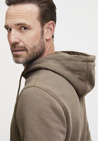Hnedá mikina s kapucňou a šnúrkou, so štruktúrovaným povrchom. Má klokanie vrecko a viditeľné švy. Model je mierne otočený s neutrálnym pozadím.