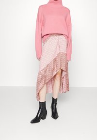 Rosa gestrickter Rollkragenpullover kombiniert mit einem gemusterten, asymmetrischen Wickelrock in hellrosa und bordeauxroten Tönen. Schwarze Knöchelstiefel runden das Outfit ab.