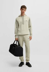 Felpa beige chiaro e pantaloni sportivi con dettagli neri, entrambi con logo. Il modello tiene in mano una borsa da viaggio nera, indossando sneakers nere.