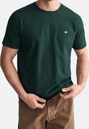 T-shirt en coton vert foncé à col rond, à manches courtes, avec un petit logo brodé sur la poitrine, associé à un pantalon beige.