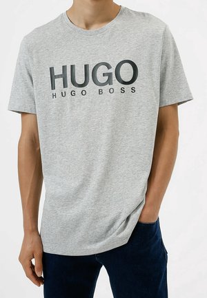 Homme portant un t-shirt à manches courtes gris clair avec un grand texte noir "HUGO" et un texte plus petit "HUGO BOSS", associé à un pantalon bleu foncé.
