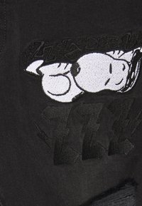 Tela de mezclilla negra con un destacado gráfico bordado en blanco de Snoopy, con costuras tonales en el fondo y elementos de texto en negrita.
