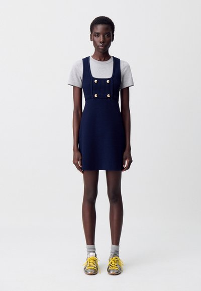 Robe mini bleu marine avec une encolure carrée, un corsage côtelé et des boutons dorés. Associée à un t-shirt gris et des baskets jaunes, complétées par des chaussettes grises.