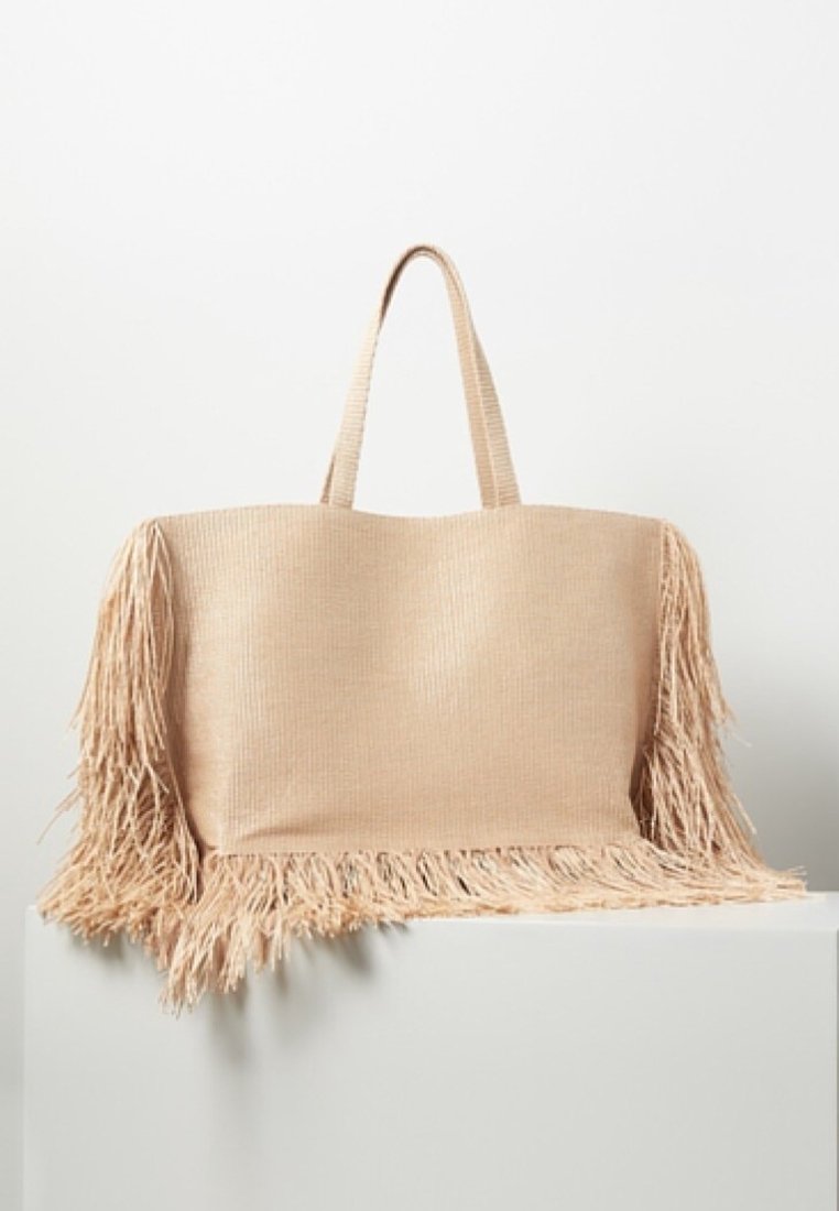 Sac fourre-tout beige en tissu texturé, doté de longs franges le long des bords et de deux poignées solides pour un transport facile.