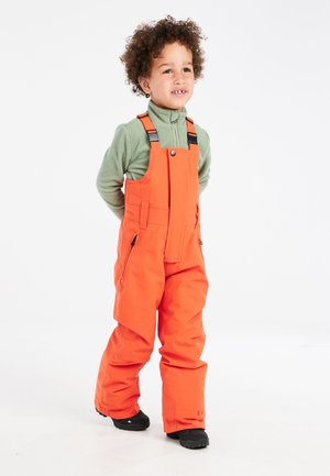 Overoles impermeables naranjas con tirantes ajustables, bolsillos laterales y una textura suave, combinados con una camiseta de forro polar verde. Botas negras.