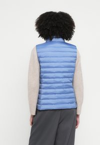 Niebieska pikowana kamizelka z wysokim kołnierzem i poziomym przeszyciem, zestawiona z beżowym swetrem. Tekstura jest gładka i puchowa.