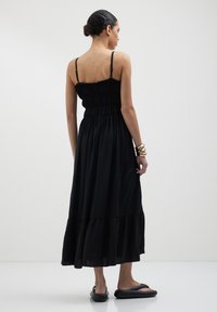 Robe maxi noire avec bretelles spaghetti réglables, corsage smocké et jupe à volants. Tissu doux avec une texture fluide.