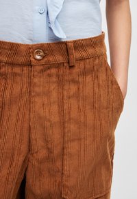 Calças de veludo cotelê castanhas com textura ribada vertical, apresentando um fecho frontal com botão e bolsos laterais, combinadas com uma camisa azul clara.