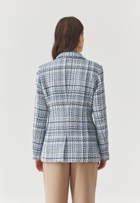 Blazer de cuadros texturizados en azul y blanco con corte entallado, mangas largas, cuello con muesca y una abertura trasera sencilla. Combinado con pantalones beige.
