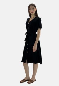 Vestito nero con bottoni, maniche corte, vita alta e cintura da annodare. Presenta una silhouette dritta e un orlo fino al ginocchio. Abbinato a sandali marroni.