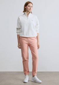 Sweatshirt branca com fecho de correr e mangas compridas, combinada com calças jogger cor de rosa claro e ténis brancos. Texturas suaves e design casual.