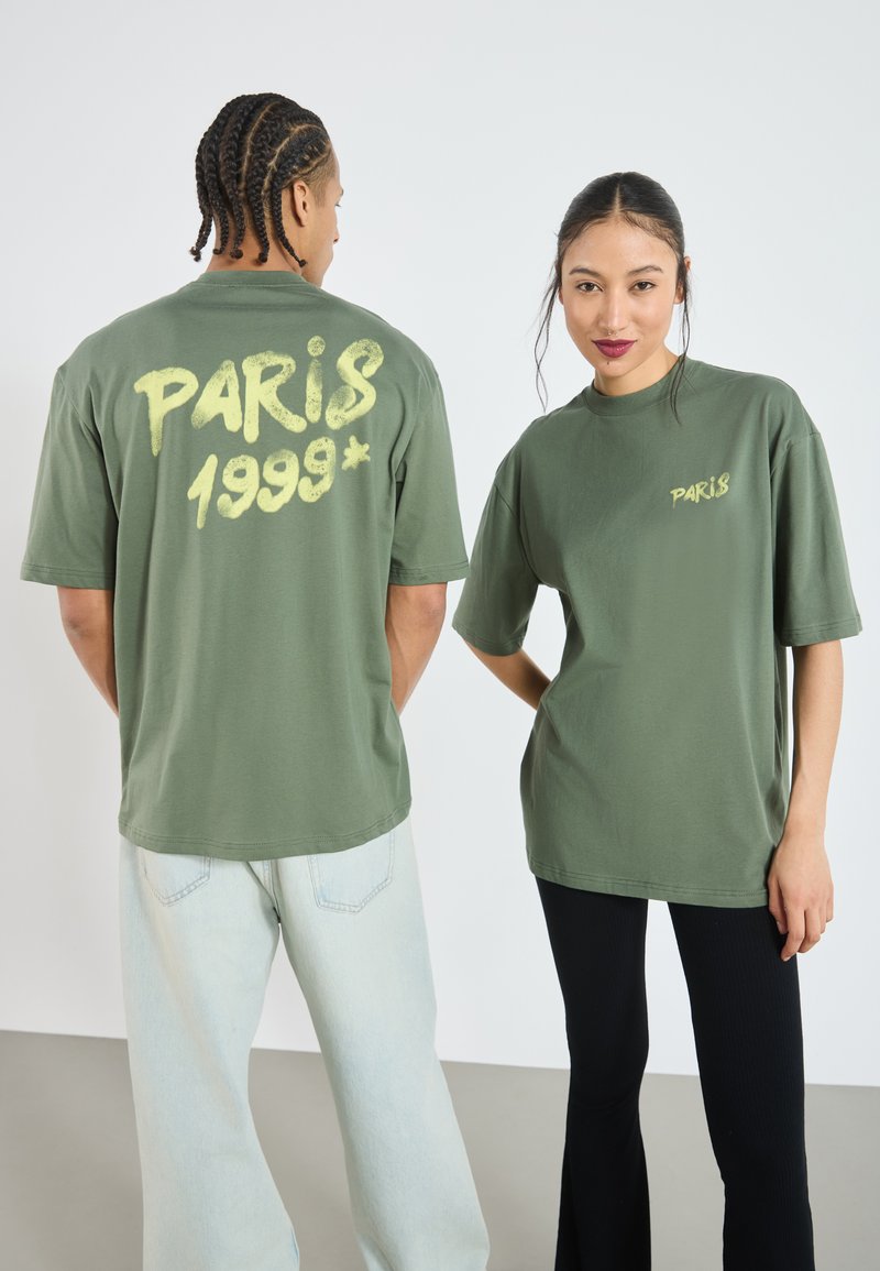 Olivengrøn oversized t-shirt med gul graffiti-stil "PARIS 1999*" tryk på ryggen og venstre bryst, båret med lyse denim og sorte bukser.