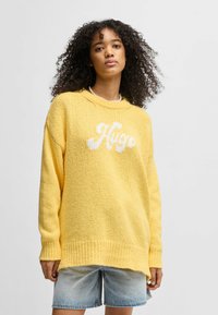 Maglione giallo lavorato a maglia con orlo e polsini a costine, caratterizzato da un design del logo bianco sul davanti. Abbinato a pantaloni corti in denim.