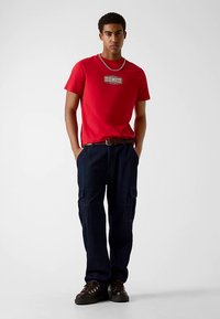 Rood katoenen T-shirt met een logopatch, gecombineerd met donkerblauwe cargobroek met zakken en bruine veterschoenen. Staande houding.