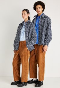 Dois modelos a usar um casaco riscado em azul-marinho e branco texturizado, uma camisa azul-clara com babados, calças de veludo cotelê castanhas e sapatos pretos robustos.