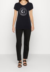Marineblå kortærmet t-shirt med rund halsudskæring, der har et centralt cirkulært logo og en glat bomuldsstruktur. Sammen med sorte slim-fit jeans og sorte sandaler.