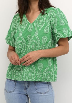 Vrouw draagt een groene geborduurde korte-mouw blouse met bloemenpatronen en lichtblauwe spijkerbroek, staand met handen lichtjes gevouwen.