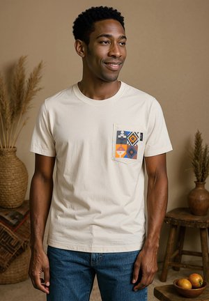 Crèmekleurig katoen T-shirt met een vierkante zak met een kleurrijk geometrisch patroon. Korte mouwen en een ronde hals. Blauwe spijkerbroek eronder.