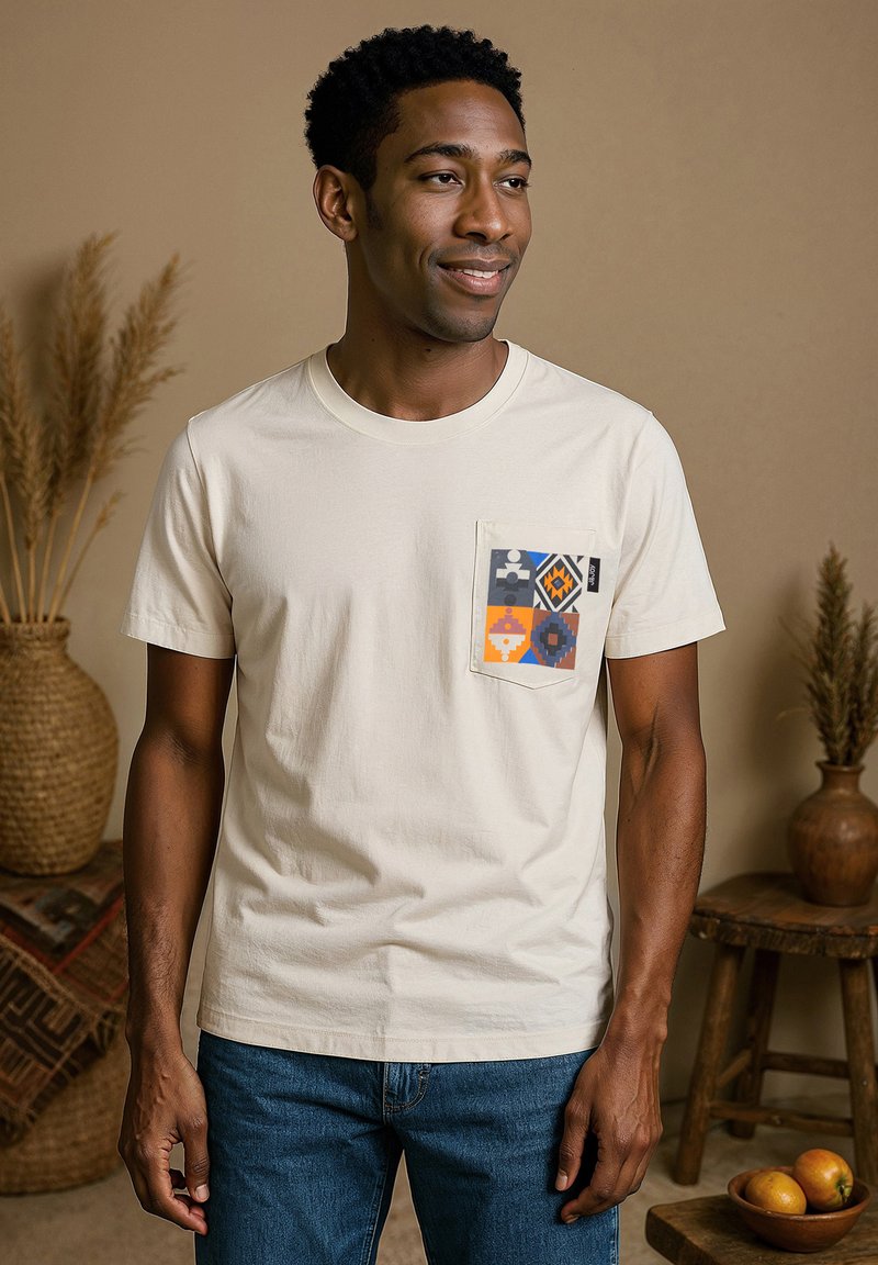 T-shirt en coton crème avec une poche carrée arborant un motif géométrique coloré. Manches courtes et col rond. Jeans bleus portés avec.