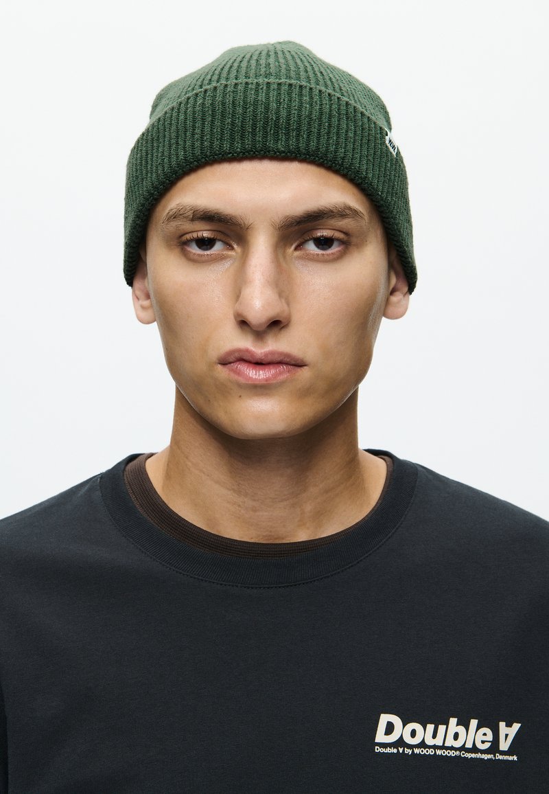 Groene geribbelde gebreide beanie met een subtiele naaddetail. Draagt een zwart T-shirt met een contrasterend logo in lichtbeige.