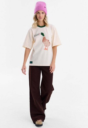 Kremowy t-shirt z zielonym dekoltem i nadrukiem kaczki. Połączony z szerokimi brązowymi spodniami i beżowymi butami. Modelka ma na sobie różową czapkę typu beanie.