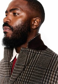 Pleds vilnas mēteli ar brūnu samta apkakli, ar augstu kaklu un mīkstu tekstūru, uzvilkts virs sarkanas krekliņas ar balto apkakli.