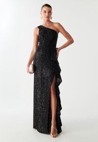 Crna haljina jednopodručnog dizajna s cirkonima po cijeloj površini, s detaljem od nabranog materijala sa strane i visokim prorezom. Uparena s malom kremasto-bijelom clutch torbicom i metalik cipelama.