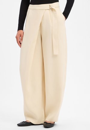Frau trägt hoch taillierte, weit geschnittene cremefarbene Hose mit Gürtel-Detail, kombiniert mit schwarzem Langarm-Top und schwarzen Ballerinas.