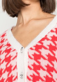 Czerwono-biały kardigan w kratę w jodełkę z dekoltem w serek, o puszystej fakturze i dwoma ozdobnymi guzikami w kształcie kropli kryształu.