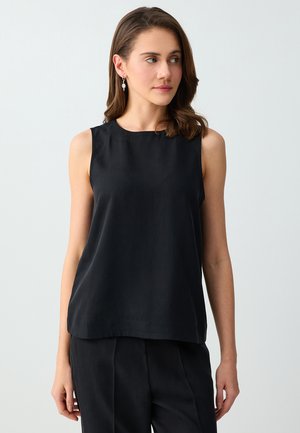 Femme portant un haut noir sans manches et un pantalon noir, regardant vers sa droite sur un fond clair uni.