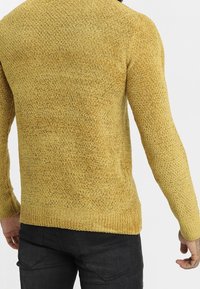 Żółty sweter o fakturze z prążkowanym brzegiem i mankietami, wykonany z gęstego splotu i dopasowany w kroju, noszony na ciemnych spodniach.