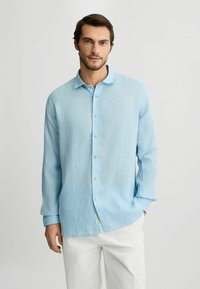 Camisa de botones azul claro en una tela suave y texturizada, con cuello clásico y mangas largas, combinada con pantalones blancos.