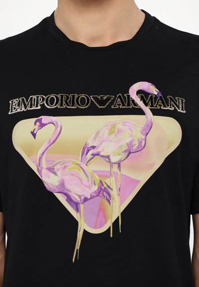 Črna majica s kratkimi rokavi, ki prikazuje grafiko dveh roza flamingov znotraj bež trikotnika, nad izvezenim besedilom "Emporio Armani."
