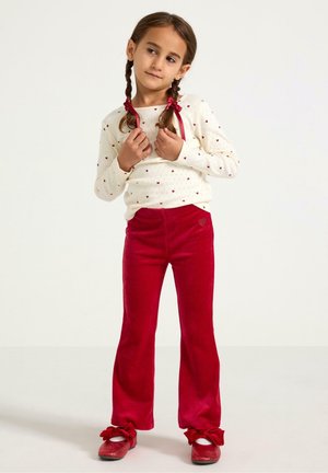 White knitted sweater with red heart pattern, paired with red velvety flared pants and red shoes featuring bows. Child posing with braids.