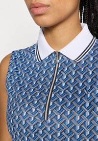 Ujjatlan polo kék és fekete geometrikus mintával, fehér gallérral és cipzárral. Kontrasztos csíkok találhatók a gallér mentén.