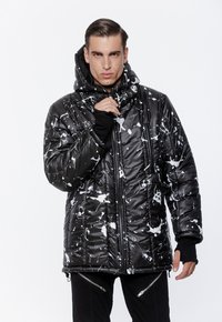 Mężczyzna w czarnej pikowanej kurtce z białymi plamami farby, zamek zamknięty, z kapturem i czarnych spodniach z detalami zamków.