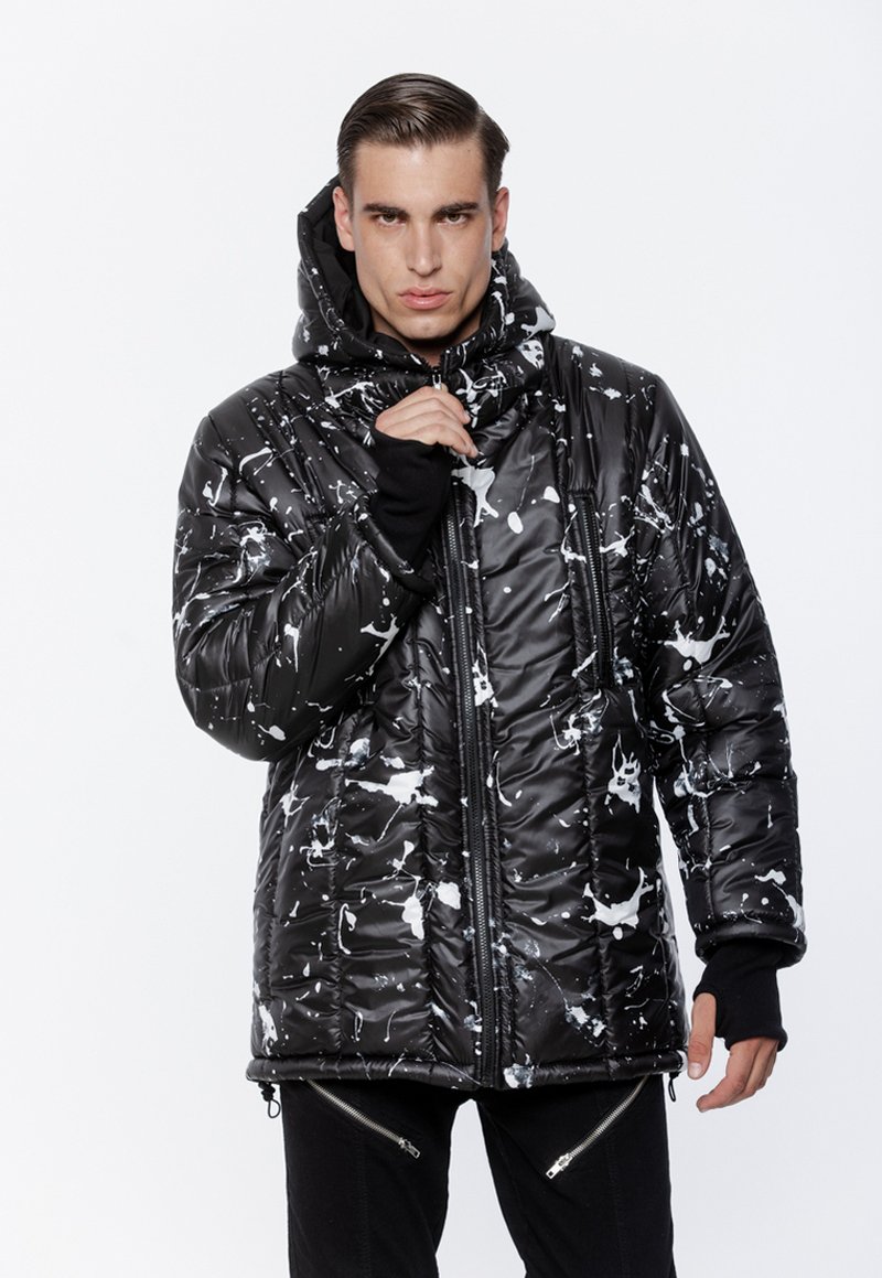 Mężczyzna w czarnej pikowanej kurtce z białymi plamami farby, zamek zamknięty, z kapturem i czarnych spodniach z detalami zamków.