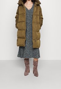 Vihreä puffer-takki hupulla, jossa on tikattu muotoilu. Sen alla tummakukkainen mekko ja polven ylle ulottuvat vaaleanruskeat saappaat, joissa on paksu korko.