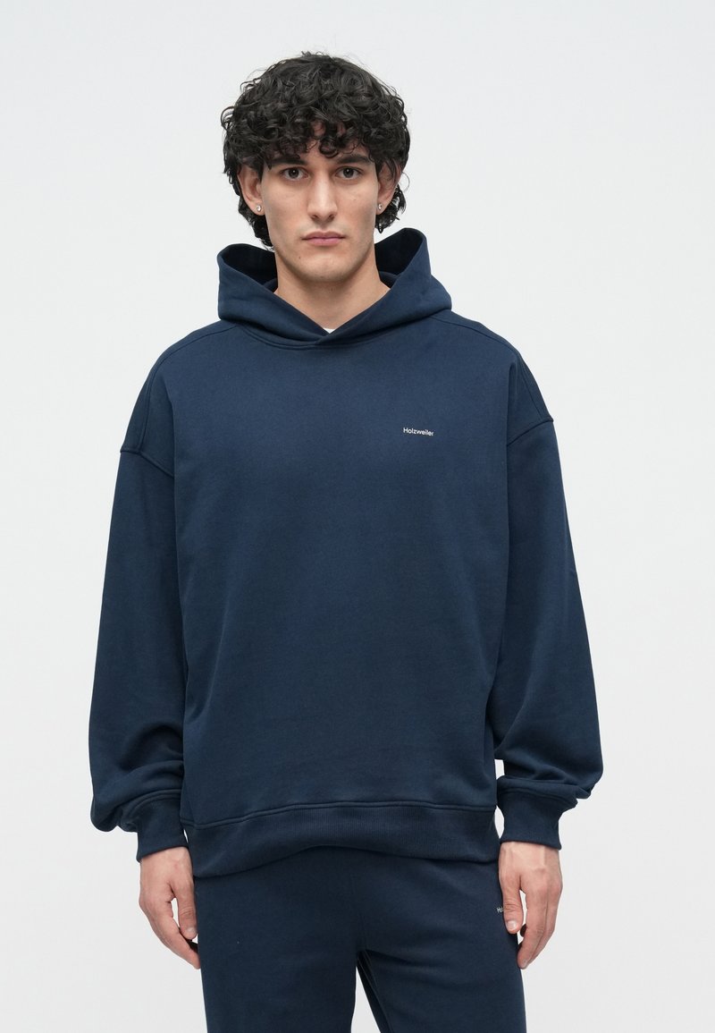 Navy blue hoodie with a drawstring hood, oversized fit, and embroidered logo on the chest. Smooth fabric and ribbed cuffs and hem.