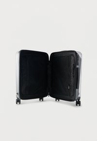 Maleta rígida plateada con cuatro ruedas, abierta para mostrar compartimentos interiores vacíos de color negro con bolsillos de malla con cremallera en ambos lados.