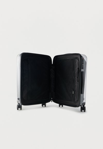 Ασημένια βαλίτσα σκληρού κελύφους με τέσσερις ρόδες, ανοιχτή για να δείχνει εσωτερικά κενά μαύρα διαμερίσματα με διχτυωτές τσέπες με φερμουάρ και στις δύο πλευρές.