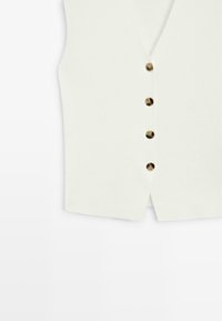Witte mouwloze bodywarmer met vier bruine knopen middenvoor en een kleine voorste split aan de zoom.