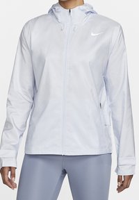 Mujer con una chaqueta con cremallera de color gris claro de Nike, con capucha y agujeros para los pulgares, combinada con leggings deportivos grises.