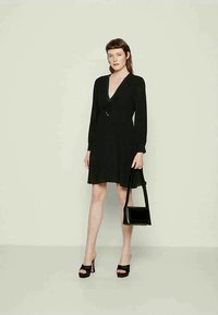 Frau trägt ein schwarzes langärmliges V-Ausschnitt-Kleid, schwarze High-Heelsandalen, hält eine schwarze Handtasche und steht vor einem hellen Hintergrund.